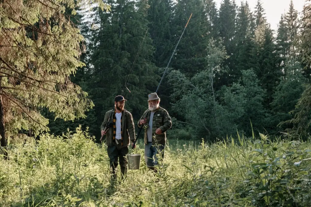 Two grown men happily talking while walking in the forest holding a fishing rod and a silver bucket