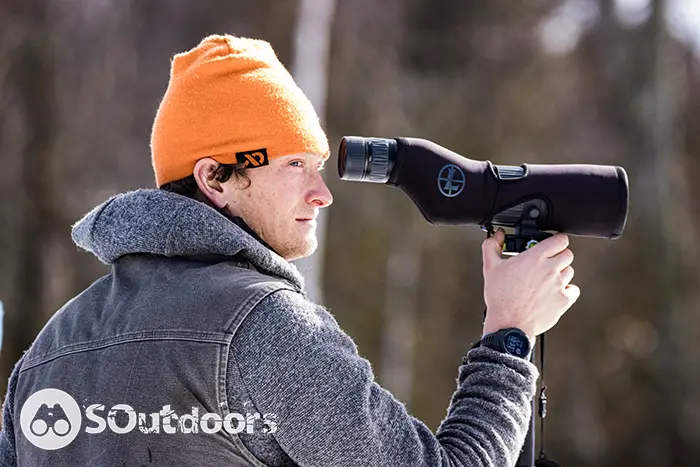 Man wearing a jacket and an orange beanie sighting on a monocular scope